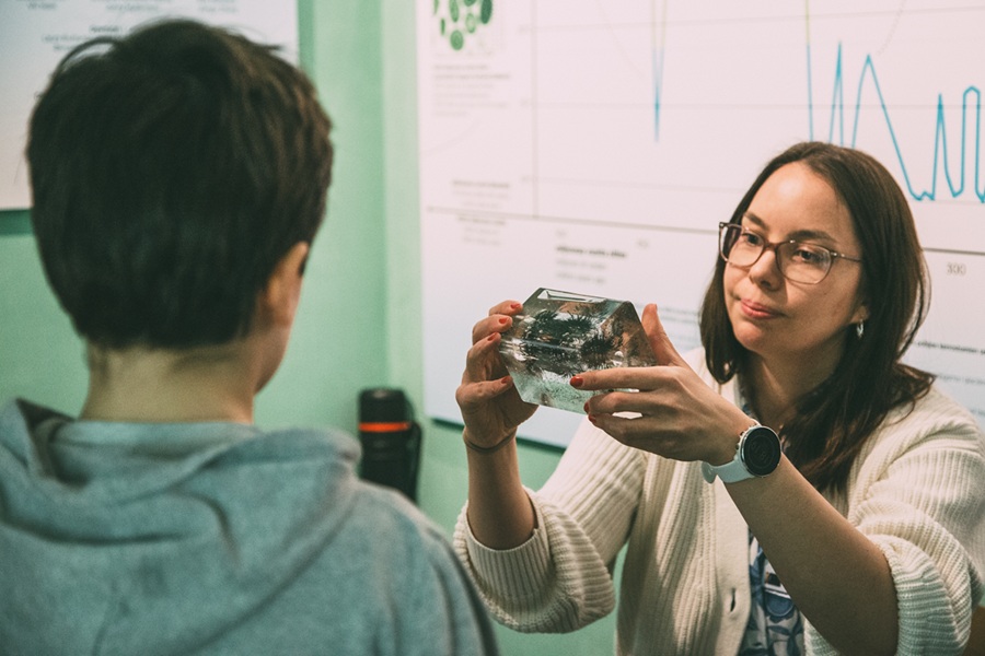 Naisoletettu henkilö pitää käsissään läpinäkyvää kuutiota, jonka sisällä on jotakin, toinen henkilö katsoo.